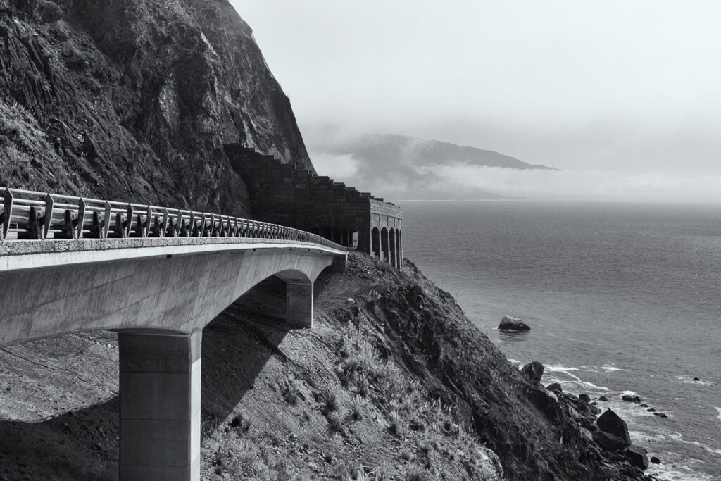 A dramatic black-and-white image of the Pacific Coast Highway in Big Sur, California, featuring a sweeping concrete bridge curving around a steep cliffside. The bridge leads to a stone tunnel with arched openings built into the rocky mountain. Below, waves crash against a rugged shoreline, and misty clouds hover over the distant coastline.