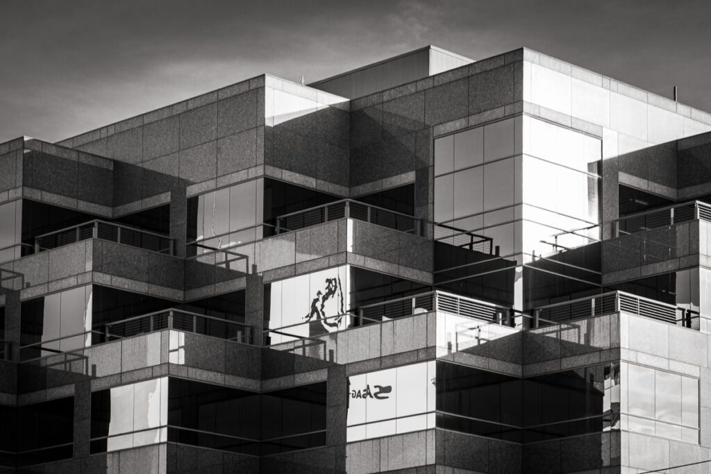 A modern glass-and-stone building in black and white, emphasizing the angular, zig-zagging design. Strong contrasts between light and shadow highlight the geometric structure, while reflections in the glass create abstract shapes. The absence of color enhances the interplay of depth, form, and mirrored distortions in the windows.